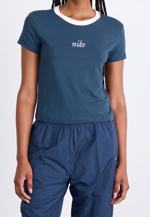 Kobieta ubrana w niebieską skróconą koszulkę Nike z białym kołnierzykiem i pasujące luźne niebieskie spodnie, stojąca na jednolitym tle.