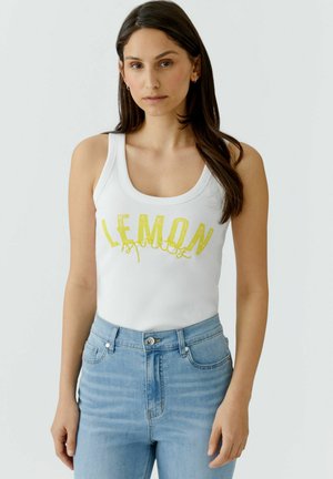Jeune femme aux longs cheveux foncés portant un débardeur blanc avec le texte jaune « LEMON » et un jean taille haute bleu clair sur fond uni.