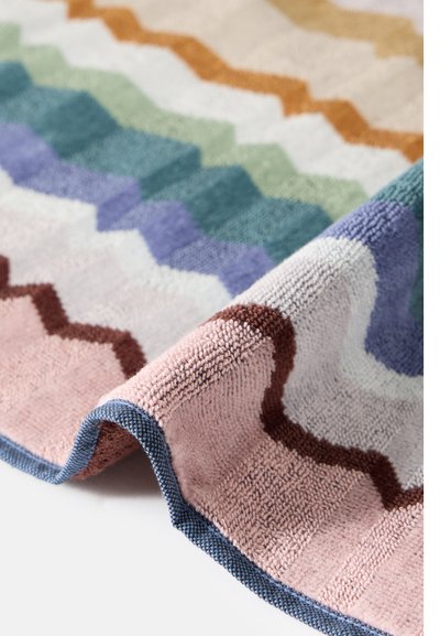 Gros plan d'un tapis doux plié avec un motif en zigzag aux couleurs pastel comprenant du rose, violet, vert et orange sur fond blanc.