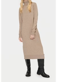 Robe à col roulé en tricot beige, avec des manches longues et des ourlets côtelés. Associée à des bottes noires montantes pour un contraste. Texture lisse sur toute la surface.