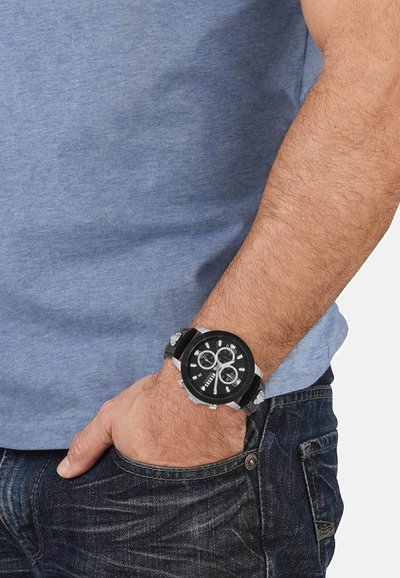 Montre noire avec accents argentés, cadran rond, trois sous-cadrans, et bracelet noir texturé. Portée sur une main reposant dans une poche.