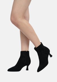 Botas de tobillo de ante negro con punta afilada, cremallera lateral y un pequeño tacón cónico transparente. Textura suave con un diseño minimalista.