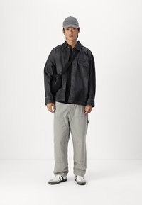 Giacca di jeans nera, top nero, pantaloni grigi e sneakers bianche con strisce nere. Cappello grigio e piccola borsa a tracolla nera. Sfondo semplice.