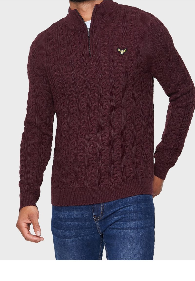 Bordeauxfarbener Strickpullover mit einem Viertelreißverschluss, der ein Kabelmuster und ein kleines gesticktes Logo auf der Brust aufweist. Getragen mit blauen Jeans.