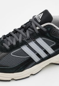 Gros plan d'une basket Adidas noire et grise avec trois bandes blanches, lacets gris, tissu en mesh et semelle rembourrée.