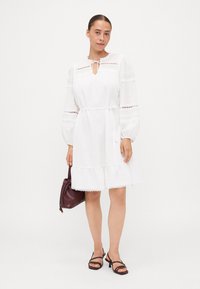 Vestido branco com mangas bufantes longas, acabamento em renda e decote com laço. Apresenta uma bainha com babados. Combinado com uma bolsa roxa escura e sandálias pretas.