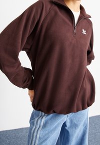 Brun fleece halvdraget tröja med långa ärmar och ribbade mudd. Har en liten vit logotyp på bröstet och en lös passform.