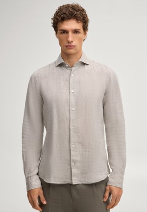 Młody mężczyzna z kręconymi włosami, ubrany w jasnobeżową, długorękawową koszulę na guziki i szare spodnie, stojący na jednolitym tle.