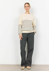 Beige svetr s černými pruhy, dlouhé rukávy, v kombinaci s tmavě šedými volnými kalhotami a světlou obuví. Jednoduchý a funkční design.