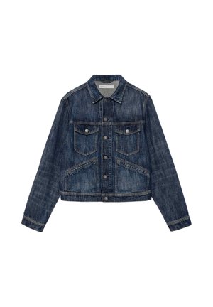 Homme portant une veste en jean bleu foncé et un jean, des bottes marron et de grandes lunettes de soleil noires, debout les mains dans les poches.