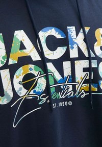 Mørkeblå hoodie med farverige, sprøjtede "JACK & JONES essentials"-tekster trykt på forsiden, inklusive "EST. 1990" med små bogstaver nedenfor.