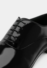Scarpe oxford in vernice nera con allacciatura chiusa e una finitura lucida e raffinata, mostrate in primo piano.