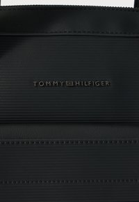 Melna Tommy Hilfiger soma ar teksturētu materiālu, reljefētu logotipu un kontrastējošām šuvju detaļām. Minimālistisks dizains ar strukturētu formu.