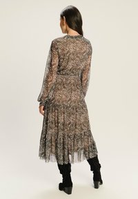 Kobieta z długimi ciemnymi włosami, ubrana w przezroczystą, wzorzystą sukienkę midi z long sleeve i czarne botki na obcasie, stojąca tyłem na neutralnym tle.