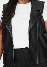 Černá kožená bezrukávová vesta s ostrým límcem, vpředu na zip a boční kapsou. Nositá přes bílý tričko. Hladká textura.