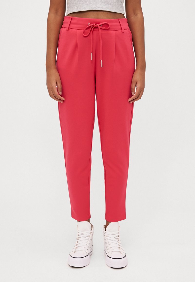 Femme portant un pantalon taille haute rose vif avec un cordon, associé à des baskets blanches montantes et un top court gris clair.