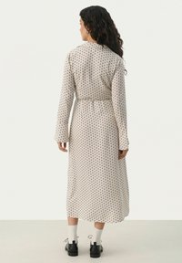 Langes, beige Kleid mit schwarzen Punkten, tailliert mit einem Gürtel. Hat lange Ärmel und einen Kragen. Wird mit schwarzen Schuhen und weißen Socken getragen.