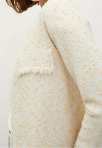 Weiche, cremefarbene Strickjacke mit strukturierten gelben Punkten und einer kleinen Vorderasche mit Fransen-Details, getragen am Oberkörper einer Person.