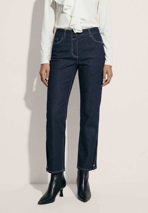 Vrouw draagt donkere rechte jeans, witte blouse met lange mouwen en ruches, en zwarte enkellaarsjes met puntige neus en hak.