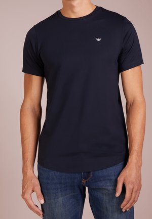 Man draagt een aansluitend effen donkerblauw T-shirt met korte mouwen en blauwe spijkerbroek, staand met ontspannen armen langs zijn zij.