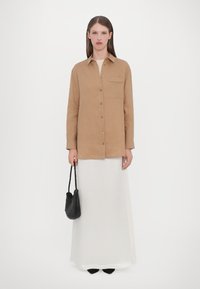 Beige long-sleeve shirt in a lightweight fabric with a pocket, button closure, paired with a white maxi skirt and a textured black bag.