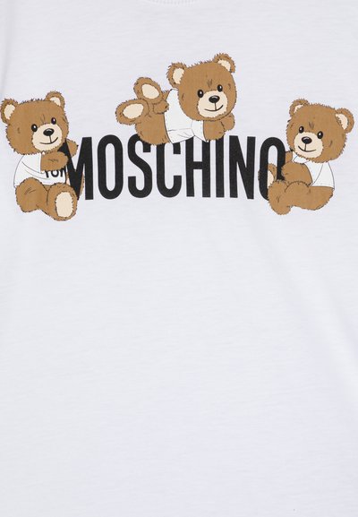 Balts T-krekls ar trim karikatūru brūnajiem lāčiem baltos krekliņos, zem kuriem ir drukāts "MOSCHINO" melniem burtiem.