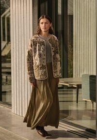 Une veste beige à motifs avec des détails brodés, un haut gris et une longue jupe olive. La tenue est complétée par des bottes marron foncé.