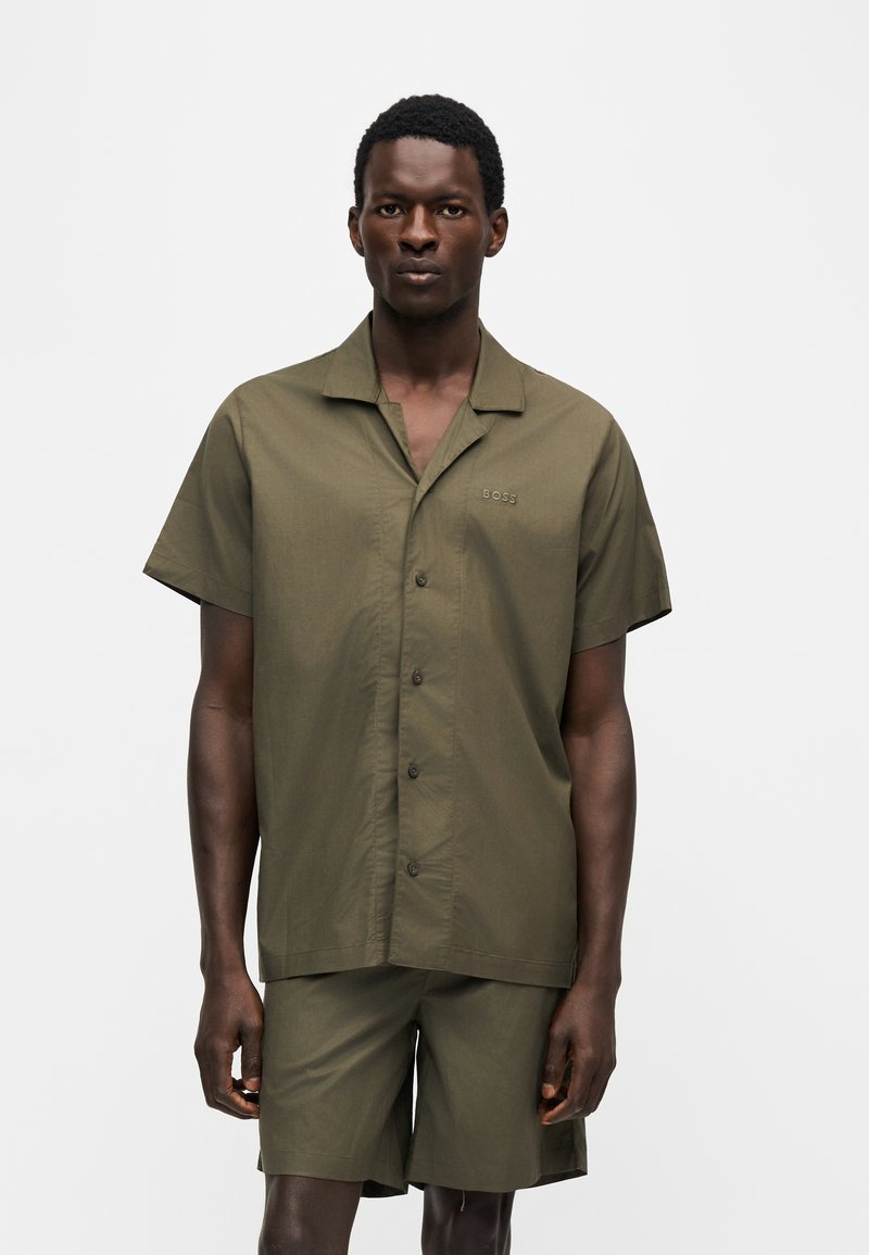 Homme debout devant un fond uni, portant une chemise olive à manches courtes boutonnée et un short assorti, face à l’objectif.