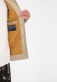 Beige jakke med lynlåsen åben, der afslører et glat, sennepsgult foer med quiltet tekstur, der har et plejelabel i den indre lomme.