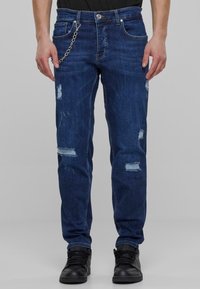 Mørkeblå denimjeans med en avslappet passform, med slitte detaljer og lyse lapper. Inkluderer en metallkjede som aksent foran.