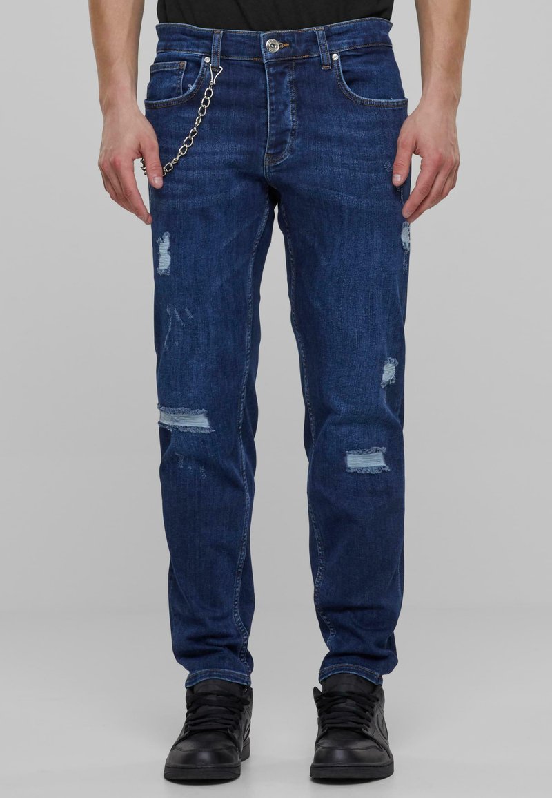 Mørkeblå denimjeans med en avslappet passform, med slitte detaljer og lyse lapper. Inkluderer en metallkjede som aksent foran.