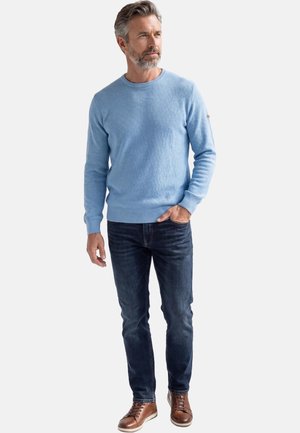 Uomo con capelli e barba grigi, che indossa un maglione azzurro chiaro, jeans scuri e sneakers in pelle marrone, in piedi con una mano in tasca.