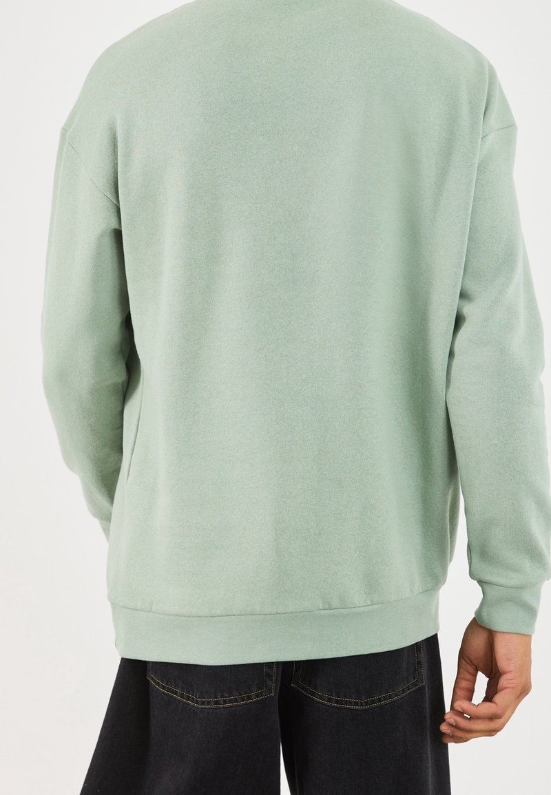 Personne portant un sweat-shirt vert clair à manches longues et un pantalon noir, vue de dos sur un fond uni.