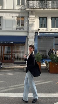 Must üleval valge särk, helesinised laiad teksad ja mustad kingad. Jalakäijate tee koos lillepotide, poesiltidega ja hoone fassaadiga taustal.