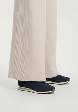 Marineblauwe sneakers met een gestructureerd bovenwerk, voorzien van een lichtgroene accenten en een bruine rubberen zool, gecombineerd met lichtbeige wijde broek.