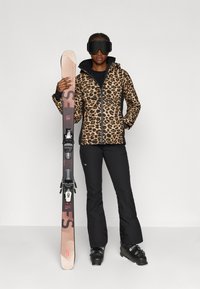 Luipaardprint jack, zwarte skibroek, houdt skies met bindingen vast; draagt skibril. Focus op textuur en kleurcontrast.