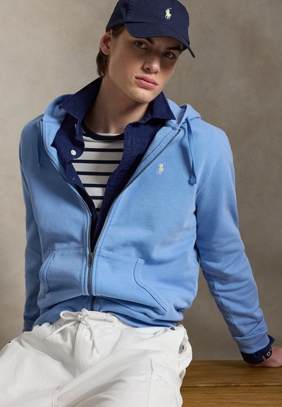 Jauns vīrietis valkā tumši zilu cepuri, gaiši zilu rāvējslēdzēja kapuci virs tumši zilas krekla un svītraina krekla, sēžot pret neitrālu fonu.