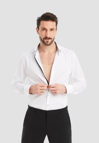 White long-sleeve shirt with a contrasting blue collar and buttons, untucked over black trousers, with hands buttoning the shirt.