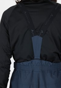 Marineblauwe overall met zwarte elastische bretels waarop de merknaam "WHISTLER" in het grijs staat. De tailleband is elastisch voor extra comfort.