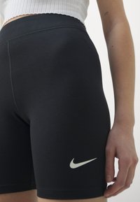 Primer plano de una persona que lleva pantalones cortos negros de Nike y un top corto blanco, mostrando el brazo con un pequeño tatuaje "777" en la parte superior del antebrazo.