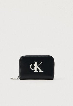 Portefeuille en cuir noir avec une fermeture éclair, arborant un logo "cK" argenté bien visible à l'avant. Surface texturée, forme rectangulaire compacte.