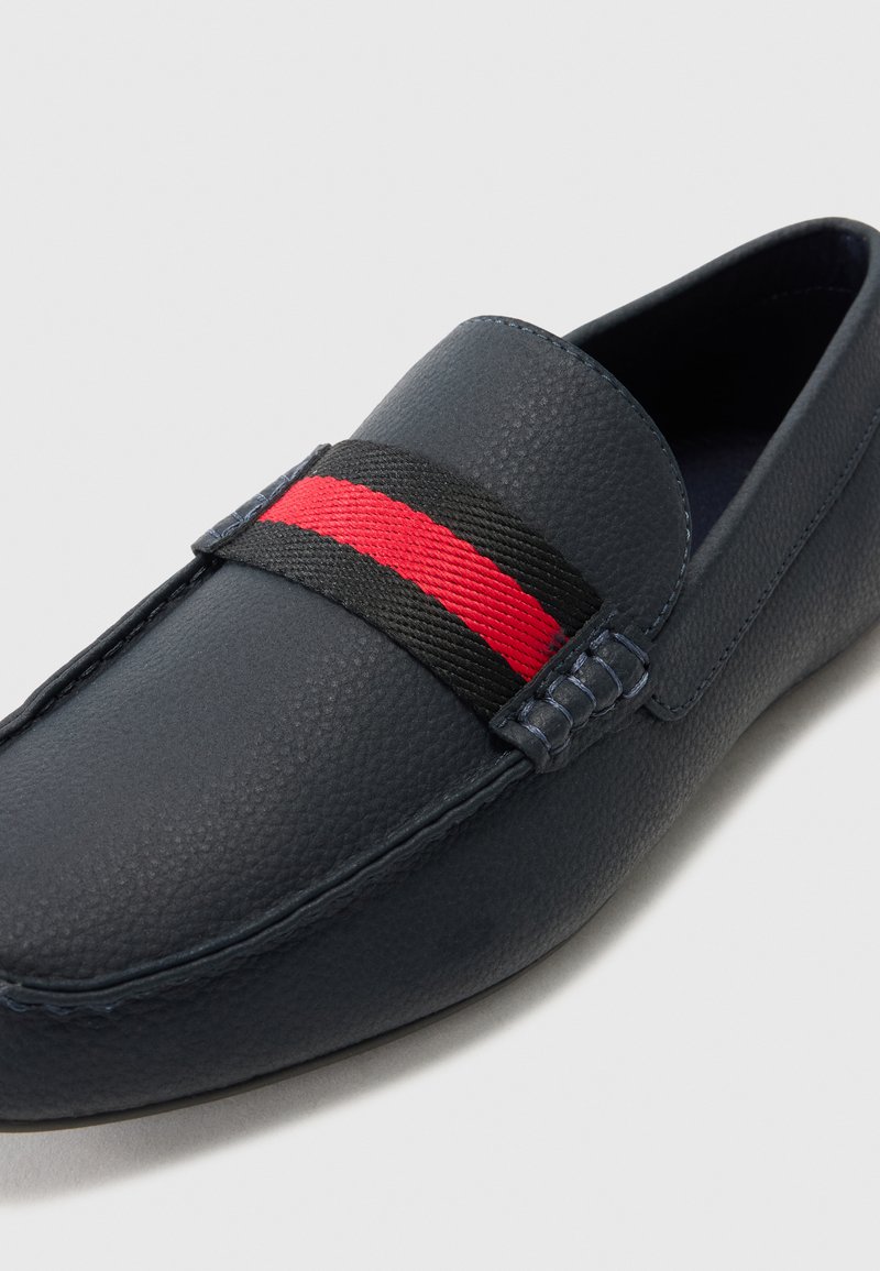 Mocasines de cuero azul marino con superficie texturizada, banda elástica a rayas negra y roja, y detalles de costura en contraste.
