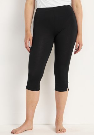 Kobieta w czarnych krótkich legginsach i białym topie, stojąca boso na jasnym, jednolitym tle.