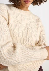 Kvinde iført en cremet kabelstrikket sweater med lange, let puffede ærmer mod en enkel hvid baggrund.