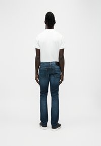 Άσπρο πόλο μπλουζάκι, μπλε τζιν παντελόνι, όρθιος από πίσω. Το τζιν έχει δύο πίσω τσέπες με αντίθετη ραφή και λείο φινίρισμα.