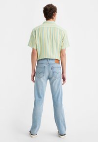 Homme portant un jean bleu clair à jambes droites et une chemise à manches courtes rayée jaune et bleue, debout dos à la caméra sur fond blanc.