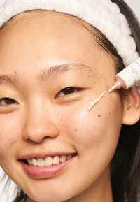 Närbild av en person som applicerar en ljusrosa kräm från en återvinningsbar tube. Slät hy med synliga fräknar och en ljus hårband.