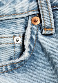 Bord effiloché de la poche d'un jean en denim bleu clair avec des coutures orange visibles et des rivets métalliques près de la passants de ceinture.