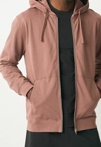 Felpa con zip di colore rosa polveroso, realizzata in tessuto morbido. Presenta tasche a marsupio, polsini a coste e cappuccio con cordino.