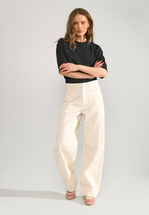 Mujer de pie con los brazos cruzados, vistiendo una camiseta negra de manga corta, pantalones blancos de cintura alta y pierna ancha, y zapatos de ballet rosas sobre fondo blanco.